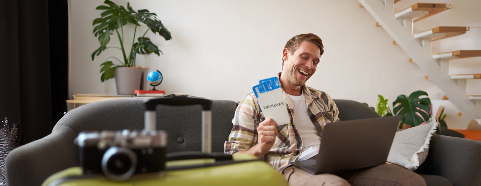 Portrait of happy tourist, man looking excited at laptop screen, holding flight tickets, going on vacation, excited about holiday, sits with suitcase in living room